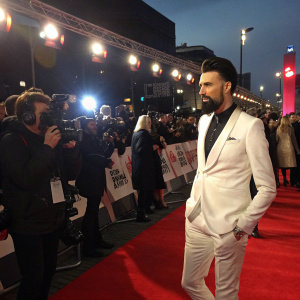 Rylan Clark-Neal, Caucasian, bearded man, dressed in a white suit, attending the BRIT Awards. Crowded red carpet scene