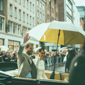 Meghan Markle and Prince Harry at an outdoor event. Smiling and waving. Beside a crowded street with tall buildings and busy pedestrians.