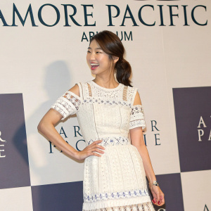 Park Shin-Hye attending the grand opening of AMORE PACIFIC 'Aritaum' in Gangnam. Wearing a white dress with intricate patterns and fringes. Cheerful expression, brown hair. Checkered background with promotional backdrop