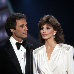 Harry Glassman and Victoria Principal at a glamorous award show. Harry in a black suit, Victoria in a white blouse. Dark, moody stage background.