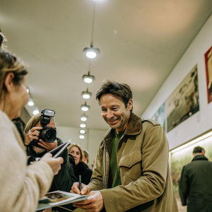 David Duchovny signing autographs. Enthusiastic fans crowd around. Bright lights and cameras flashing.