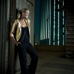 Carrie Underwood, at the Grammy Awards. Stylish outfit, black pantsuit with a gold blazer. Hair in a sleek ponytail. Posing confidently with a radiant smile, the backdrop is a bright, neon-lit cityscape.