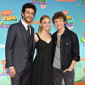 Fred, Sophia, and Joe are attending the Nickelodeon Kids' Choice Awards. Fred is dressed in a formal dark gray suit. Sophia is wearing a black dress. Joe is in a casual yet stylish outfit. The vibrant backdrop is filled with the Nickelodeon logo.