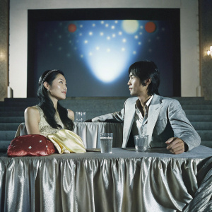 Shu QI and Chen Kun at the Cannes Film Festival. Shu QI with a long, dark hairstyle, Chen Kun with neat hairstyle. They are seated at a luxurious table with glass of water and nameplates. Behind them, a vibrant starry night projection.