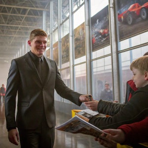 Max Verstappen in a sleek, black suit, signing autographs at an indoor fan meet. Cheerful expression