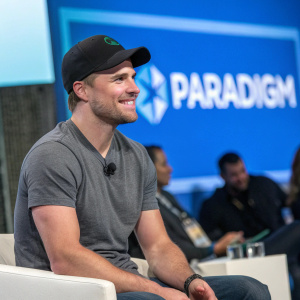 Stephen Amell, in a casual outfit, at a tech conference. Gray t-shirt, black cap, smiling. Blue Paradigm logo on backdrop.