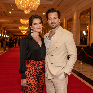 At a glamorous awards show, Nichole Gustafson and Giacomo Gianniotti make a stunning appearance. Nichole is clad in a chic black blouse paired with a vibrant leopard print skirt, while Giacomo sports a sophisticated light beige suit.