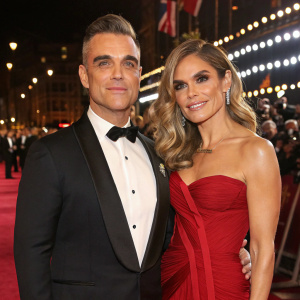 Robbie Williams and Ayda Field at a glamorous award show. Both dressed in elegant evening wear. Flashing lights and crowds in the background.