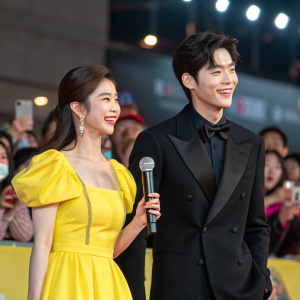 Shin Se-Kyung and Cha Eun-Woo, stars of 'Rookie Historian Goo Hae-Ryung', at a high-energy fan meet. Shin wears a vibrant yellow dress. Cha sports a sleek black tuxedo. Lively audience cheers in the background.