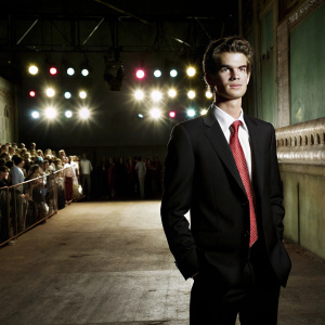 Andrew Garfield standing at the award show. Wearing a black suit with red tie. The background is a bustling crowd with colorful spotlights