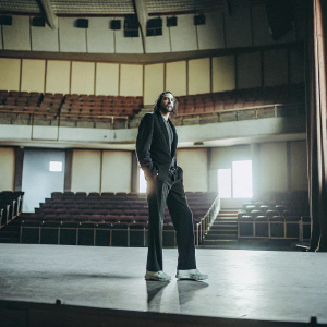 Colin Kaepernick in a sleek black suit, standing alone on an empty stage.
