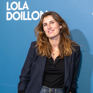 Lola Doillon, French actress, in casual attire. Wavy chestnut hair, warm smile. Dark blazer, dark top. Solid blue background with white text.