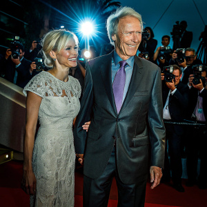 Clint Eastwood and Sienna Miller at Cannes Film Festival. Sienna Miller in a sheer white gown with intricate lace detailing. Eastwood in a charcoal suit and a lavender tie. Both smiling for the cameras on a bustling red carpet.