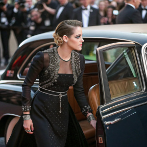 Kristen Stewart at the Met Gala, wearing an elegant black dress with intricate beadwork. Stepping out of a vintage Rolls Royce, crowd visible
