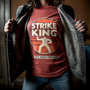 Casual photo of a red t-shirt featuring 'Strike King' logo and the phrase 'Let's Knock Them Down'. The design includes a stylized graphic of a figure in a dynamic pose.
