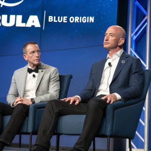 Jeff Bezos and Tory Bruno, seated together at a press conference. Elegant attire and serious expressions. Background displays ULA and Blue Origin logos on a sleek blue backdrop.