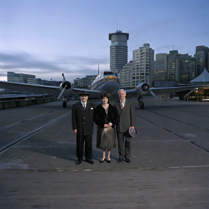 Arturo Toscanini, Carla De Martini, and Walter Toscanini, dressed formally, posing next to a Trans World Air Lines aircraft. Modern urban backdrop.