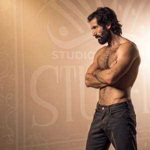 A rugged man with a muscular build, dark hair, and a beard. Posing uncomfortably in a studio. Warm, muted color tones. Background features a gradient design with detailed text.