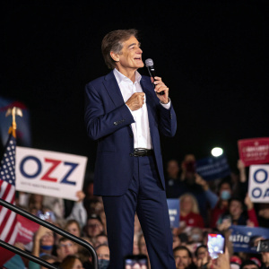Dr. Mehmet Oz speaking at a campaign rally. He wears a navy suit and white shirt. A sea of supporters in the background.
