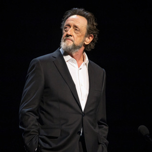 Jonathan Banks, actor, standing alone on stage at award show. Dark hair, full beard, black suit, white shirt, against solid black background.