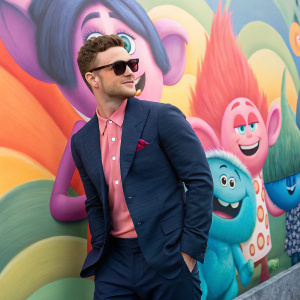 Justin Timberlake in promotional photoshoot for Trolls movie. Poses with vibrant, cartoonish Trolls imagery in the background. Wears a dark blue suit with pink shirt underneath and sunglasses.