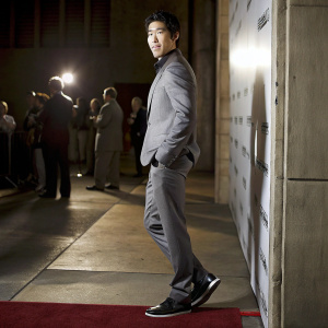Jon Lee Brody, at the premiere of Furious 7 in Hollywood. East Asian ethnicity, dark skin, wearing a gray suit, black sneakers. Confident pose against white backdrop with Furious 7 logo. Formal event, red carpet setting, other guests present.