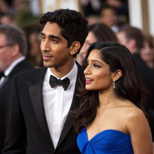 Dev Patel and Freida Pinto at the Golden Globe Awards. Both are dressed in formal attire. Dev in black tuxedo, Freida in a stunning blue gown. Crowded room, blurry faces in background
