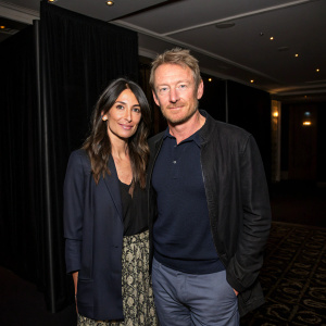 Richard Roxburgh and Silvia Colloca at an elegant evening event. Wearing casual outfits. Posed in a dimly lit room, against a black backdrop.