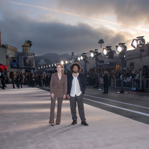 Juliette Menke and Salvatore Greco at a bustling Hollywood premiere. Juliette in a brown blazer, Salvatore in a crisp white shirt. Flashing cameras and crowd in background.