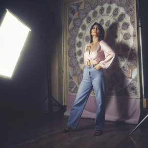 Dulce Maria, a vibrant Latina, in a photo shoot. Wearing a pastel pink blouse and blue jeans. Standing confidently in front of a large, colorful mural. Studio setting, wooden floor, high contrast lighting.
