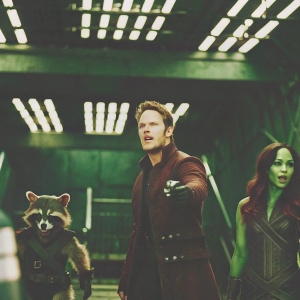Chris Pratt, Zoe Saldana, and Rocket Raccoon in an action scene from Guardians of the Galaxy. They stand in a high-tech, dimly lit factory with neon lights. Industrial machinery and control panels are visible in the background.