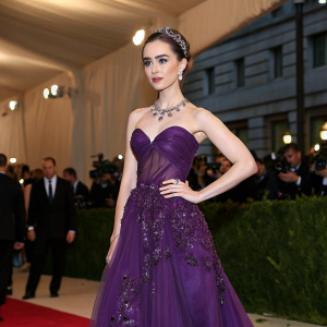 Lily Collins at the Met Gala. Wearing a stunning deep purple gown with intricate beadwork. The backdrop features the iconic Met Museum exterior.