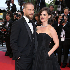 Tom Hardy and Charlotte Riley at Cannes Film Festival. Tom in black tuxedo, Charlotte in elegant black gown. Festive atmosphere with red carpet and flashing cameras.