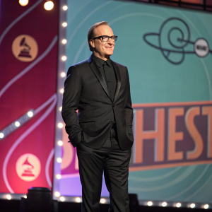 Bob Odenkirk, wearing a sleek black suit and glasses, stands confidently on a stage bathed in the soft glow of stage lights. Behind him, a vibrant, colorful backdrop with bold logos.