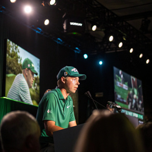 Matt Fitzpatrick, in a green polo shirt and 'Workday' cap, addressing a packed press conference. The stage is dimly lit with spotlights highlighting him. Large screens display golf tournament footage behind him.