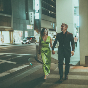 Daniel Craig and Satsuki Mitchell, both dressed to impress, walking down a bustling city street at night. Daniel in a sleek black suit, Satsuki in a vibrant green evening gown.