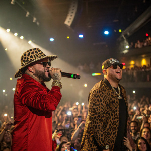 Farruko and El Alfa performing at a packed concert venue. Farruko sports a leopard-print hat, while El Alfa wears sunglasses.