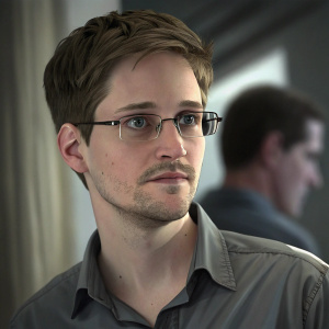 Edward Snowden, wearing glasses and a gray shirt, in a candid close-up. Background blurred, another person partially visible.