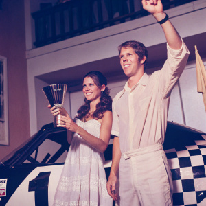 Brad Keselowski and Rachel Rupert celebrating their NASCAR win. The black and white racing car in the background, surrounded by cheering crowd.