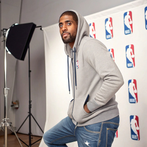 Kyrie Irving, wearing a casual hoodie and jeans, posing for a headshot. Studio backdrop with NBA logos