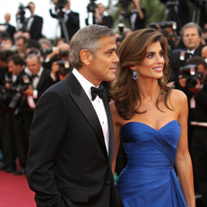 George Clooney and Elisabetta Canalis at the Cannes Film Festival. Clooney, balding, in a black tuxedo, while Canalis dons a strapless blue gown. Crowded red carpet with flashing cameras.