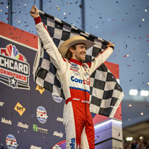 Ryan Blaney celebrating his NASCAR win. Wearing a cowboy hat and racing suit. Standing on a stage surrounded by sponsor logos, confetti, and a large checkered flag.
