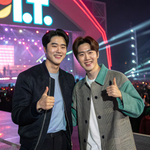 Suho and Se Hun from EXO, thumbs up pose, smiling. Bright, colorful backdrop with i.t. logo. Concert event.