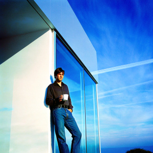 Fran Kranz, as Topher Brink, stands in front of a sleek modern office. Dressed in a dark shirt and jeans. He holds a coffee mug and leans against a glass window, looking casual yet professional