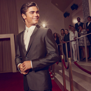 Zac Efron, wearing a sleek black suit and white shirt, at a glamorous awards ceremony. Crowd in the background blurred out