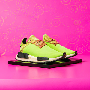 A pair of Adidas NMD TR sneakers with Dragon Ball Z theme. Shown on a sleek black platform against a backdrop with Asian-inspired scrollwork. Mostly green with gold detailing, featuring a dragon pattern. Modern style with grid-patterned sole and lace-up design. Sporty and contemporary.