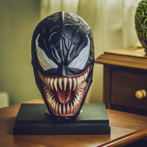 Concept art of the Venom mask from Marvel Comics. Positioned on a sleek black platform. The mask features intricate gray designs and menacing open mouth with sharp teeth. Set against a minimalist gray backdrop