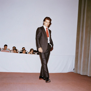 Tom Cruise, intense expression, dressed in a black suit with red tie. Studio audience in the background.