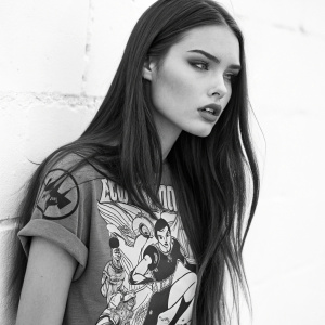 Monochrome portrait of a young woman with long dark hair in a sleek, straight style. She wears a graphic t-shirt featuring animated characters in action poses. Against a stark white backdrop, she has a serious demeanor.