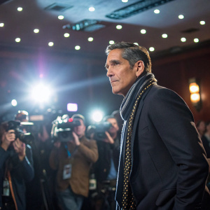 Jim Caviezel, in a dark suit and stylish scarf, poses intensely at the press conference. Bright lights and camera flashes fill the room.